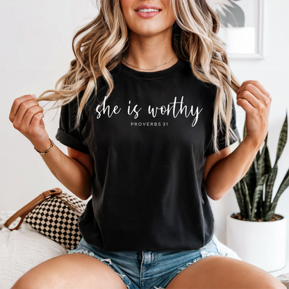 Woman wearing a black t-shirt with 'she is worthy' text, sitting on a couch.