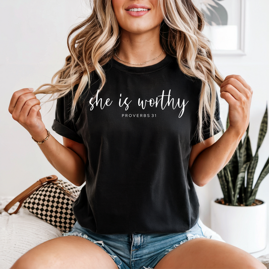Woman wearing a black t-shirt with 'she is worthy' text, sitting on a couch.
