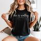 Woman wearing a black t-shirt with 'she is worthy' text, sitting on a couch.